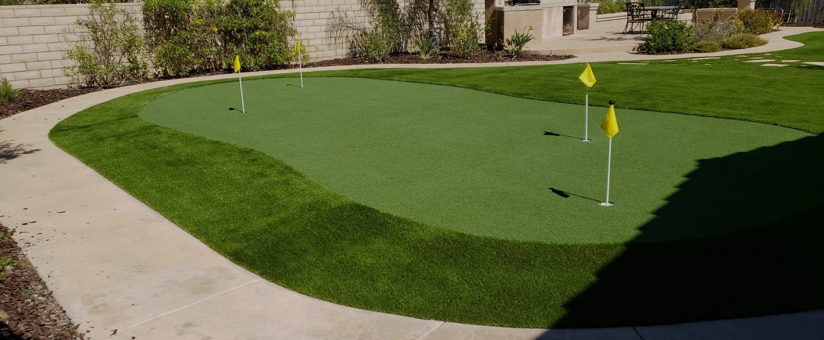 Green putting green with two yellow flags, surrounded by landscaped grass and concrete paths