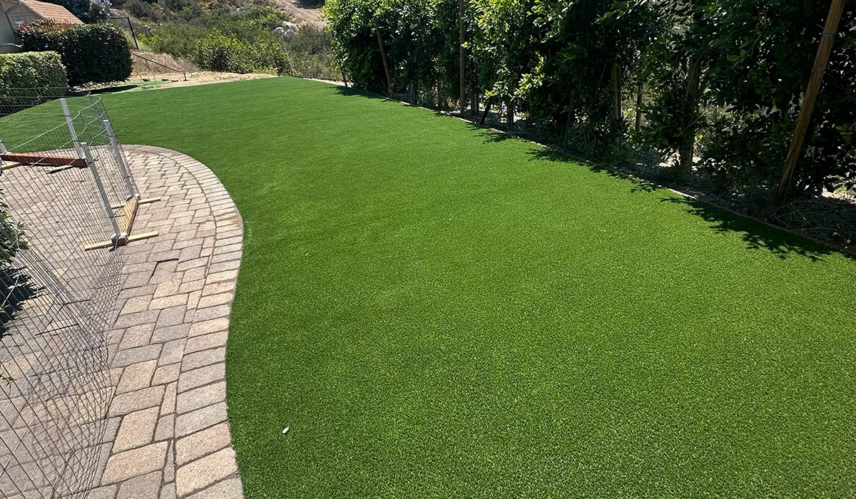 A green synthetic lawn with a curved stone pathway beside a fence and trees