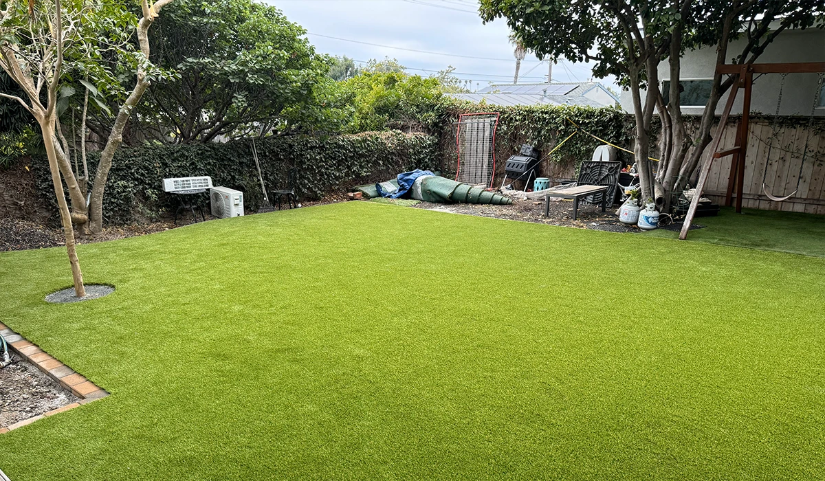 Lawn area with artificial grass, surrounded by trees and a fence. Tools and supplies in the backgrou