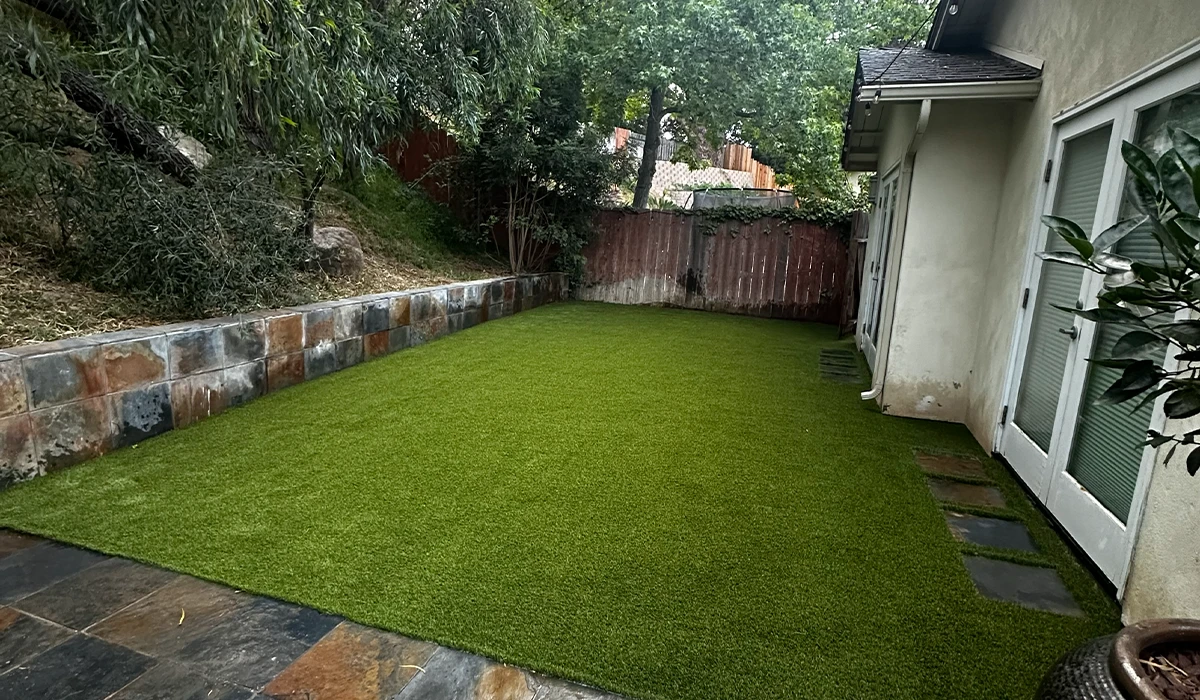Lawn area with artificial grass and stone pathway next to a house, surrounded by greenery