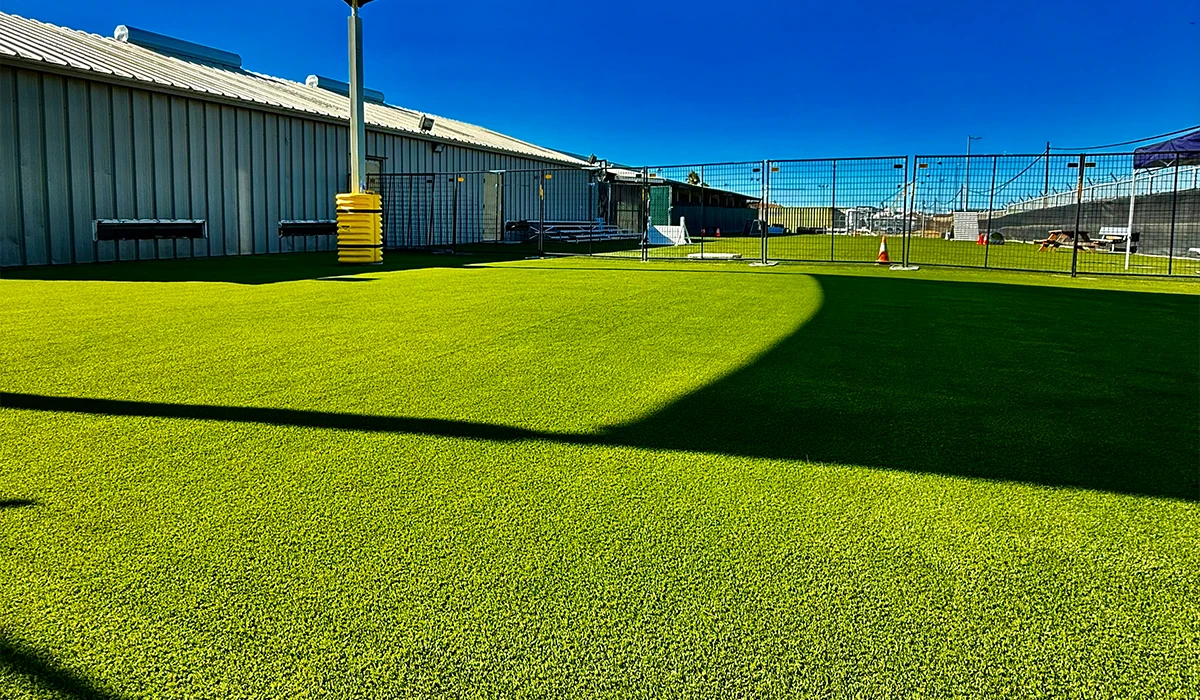 Green artificial turf with a shadow, beside a building and fenced area. Clear blue sky