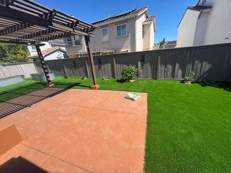 Backyard with synthetic grass, patio area, and surrounding fence. Two houses in background