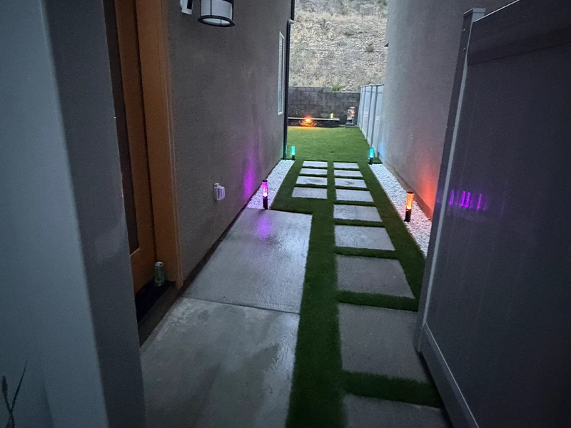 Narrow pathway with concrete slabs, grass, and colored lights on either side