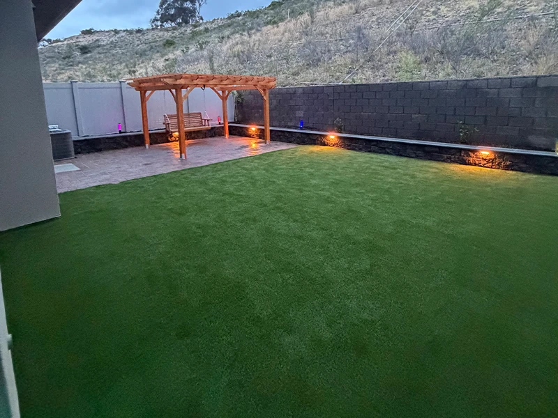 A backyard with artificial grass, a wooden pergola, and low landscape lighting