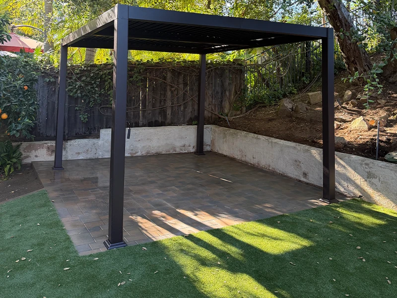 Black metal gazebo with a paved floor and green grass, surrounded by trees and a wooden fence